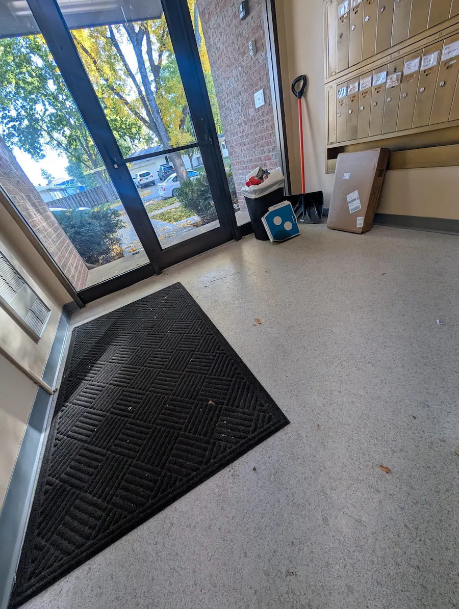 Apartment building entryway and mail area in Fargo, ND before commercial cleaning, showing tracked-in dirt, scattered leaves, litter, and a displaced floor mat